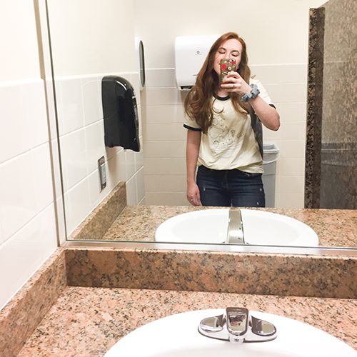 A woman stands in front of a public bathroom mirror taking a selfie. The sink has red marble around it.