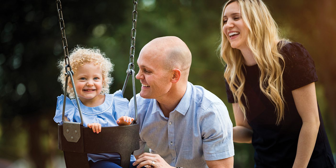 All three with smiles, an attentive father kneels down next to his daughter who is seated in a playground swing as a mother observes.