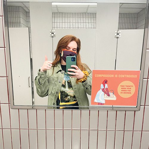 A woman stands in front of a public bathroom mirror taking a selfie.