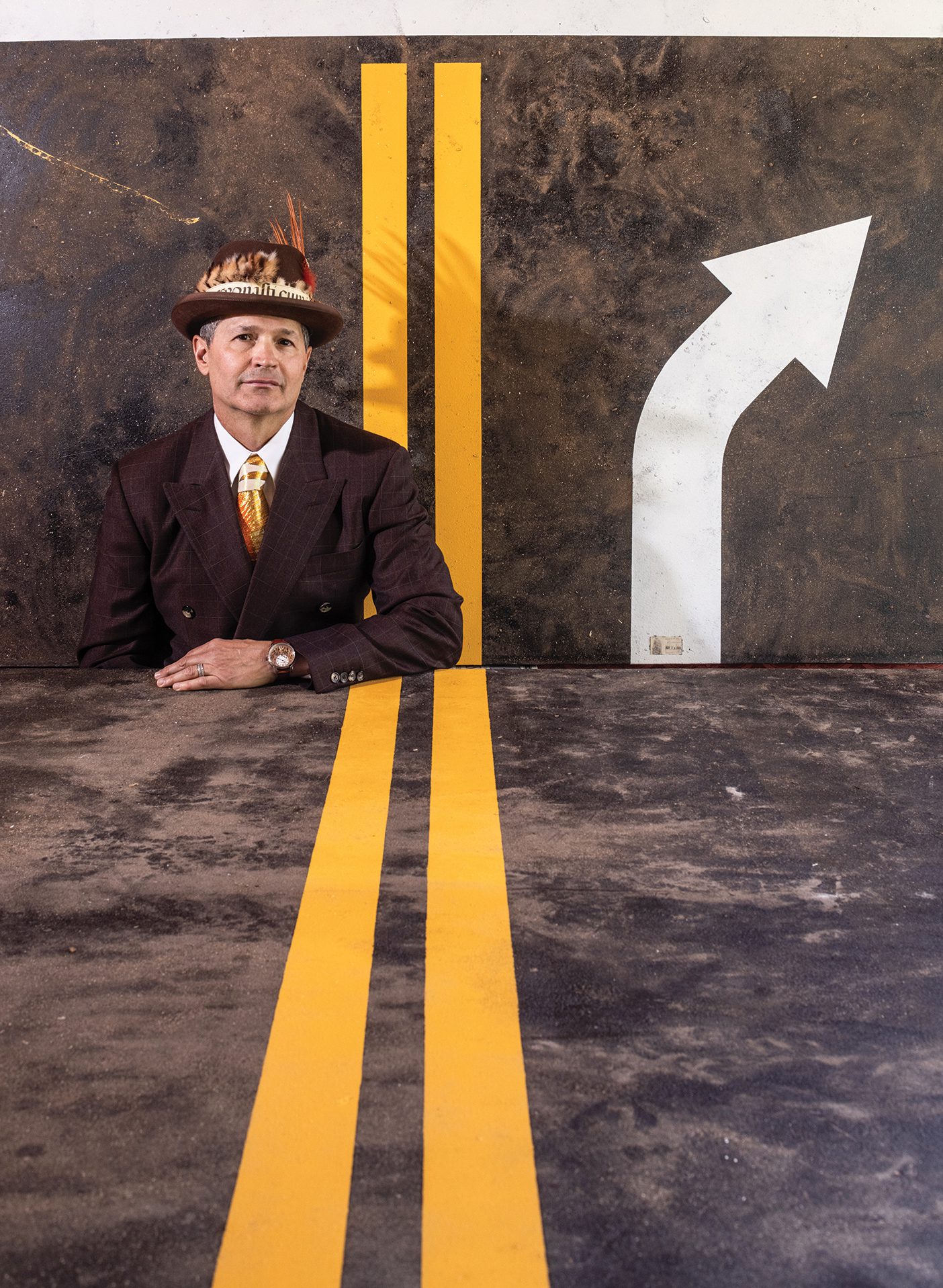 Artist Todd Stilson, wearing an orange tie and feathered top hat, stands in front of his road-inspired artwork. A table, also painted to look like a road, covers the bottom half of his body.