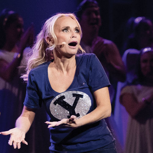A blonde woman, Kristin Chenoweth, sings onstage while wearing a sparkly BYU shirt.
