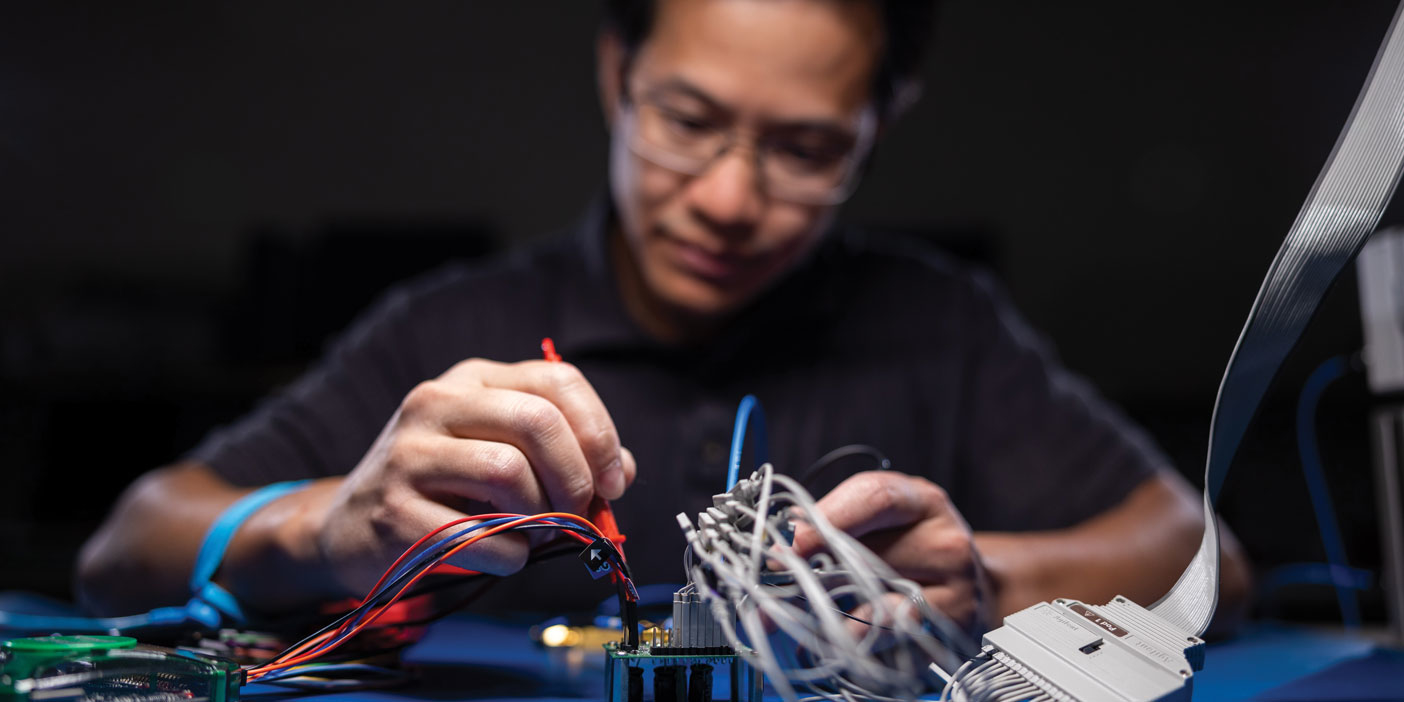 A man in glasses uses tools to adjust red, blue, and gray wires going into a microchip.