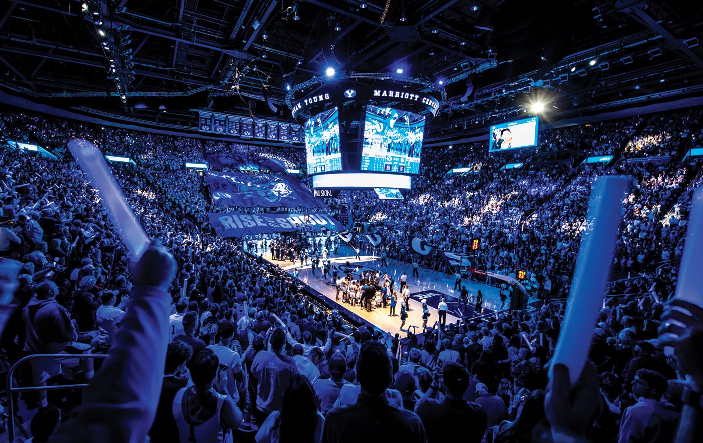 A packed Marriott center is cheering at a BYU football game. The lights are low and a "Rise and Shout" banner is held across the crowd.