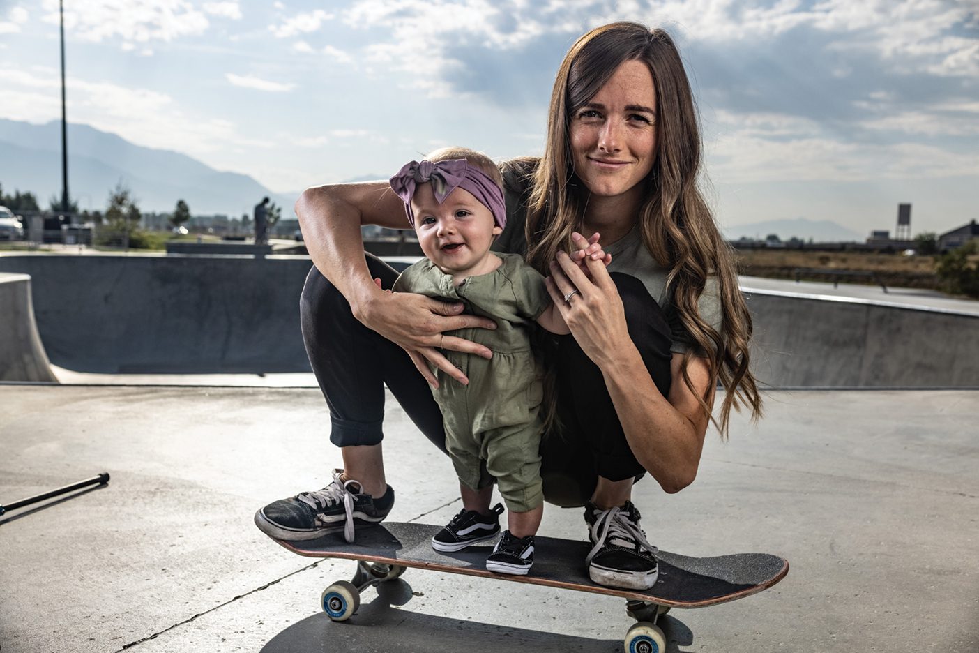 A photo of a mom crouching on her skateboard while helping her toddler daughter balance on the board.