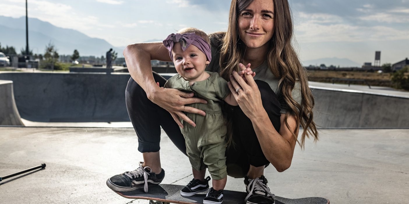 A photo of a mom crouching on her skateboard while helping her toddler daughter balance on the board.