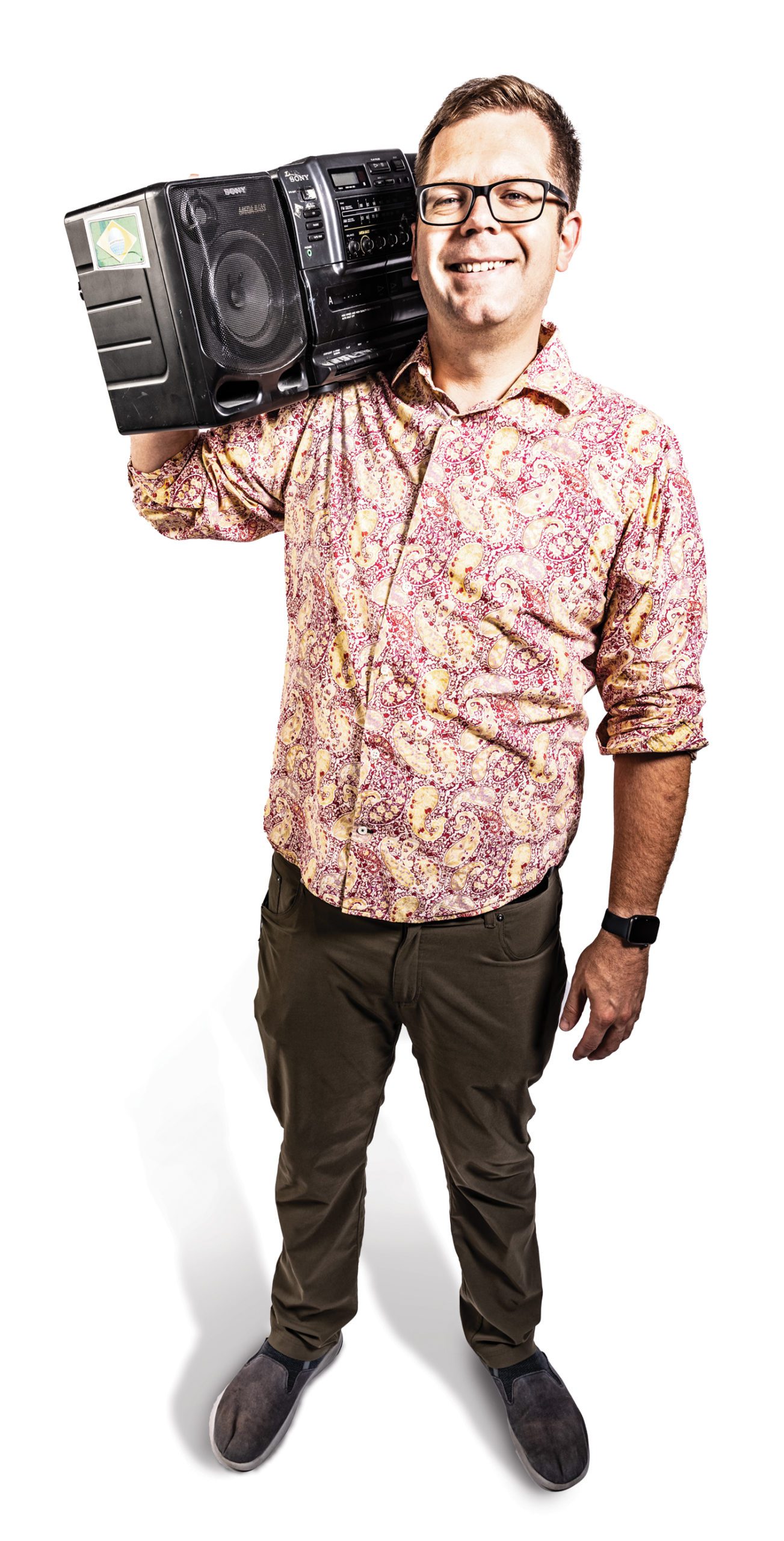 A photo of a man with black glasses and a paisley shirt balances a boom box on his shoulder.