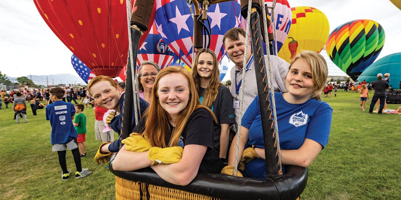 A photo of a family smiling from inside their hot-air balloon. Other balloons surround them.