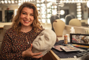A woman in a makeup studio lit with incandescent bulbs holds a white live cast of Tom Holmoe's face.