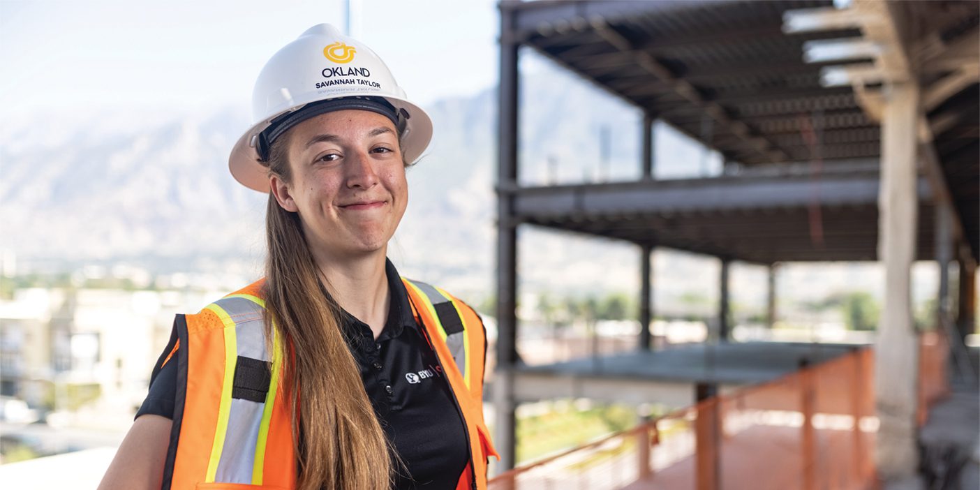 A young woman smiles while wearing a construction hat and orange vest. She stands amidst scaffolding.