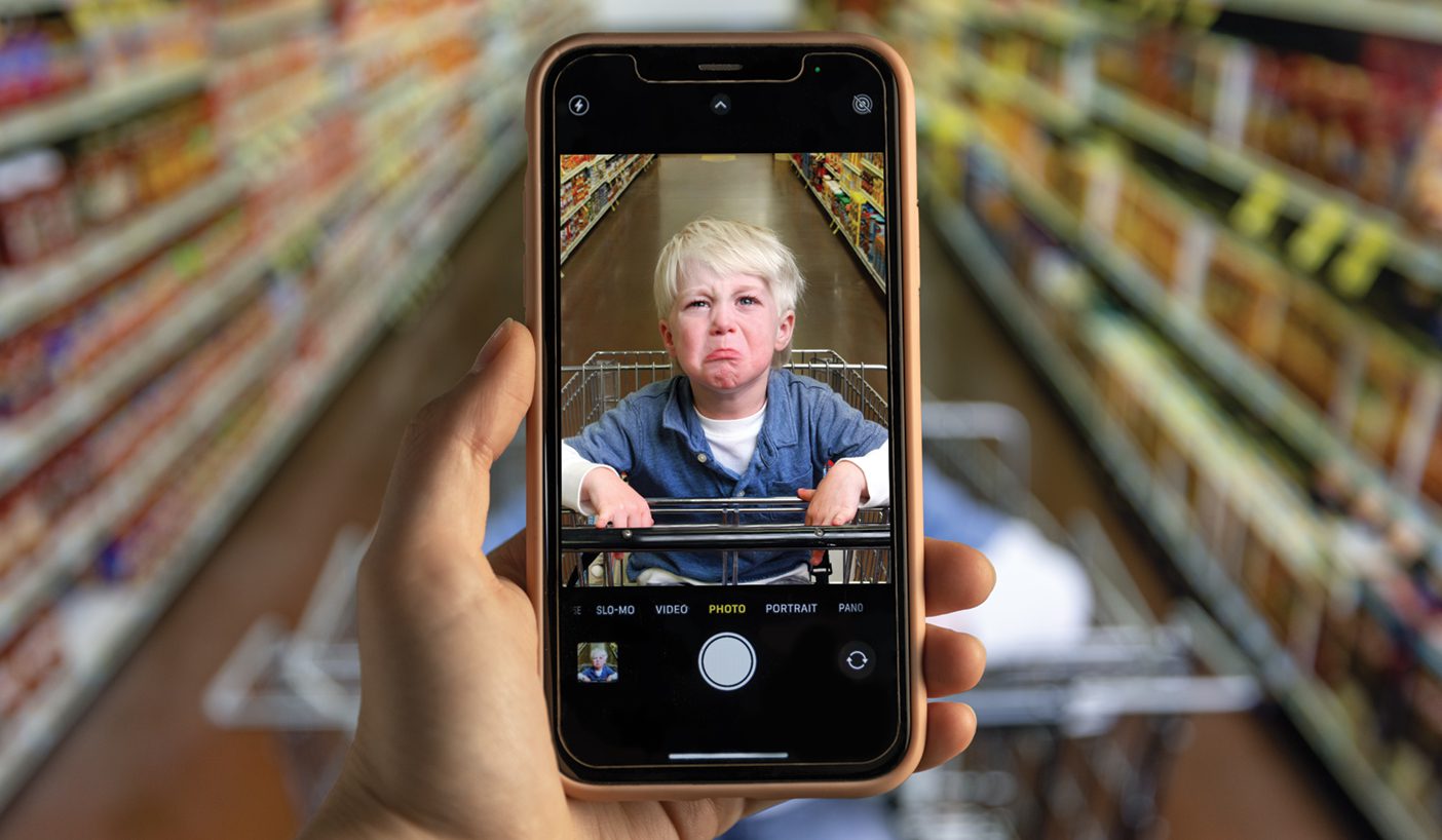 A phone zooms in on a grumpy toddler sitting in a cart in the grocery store.