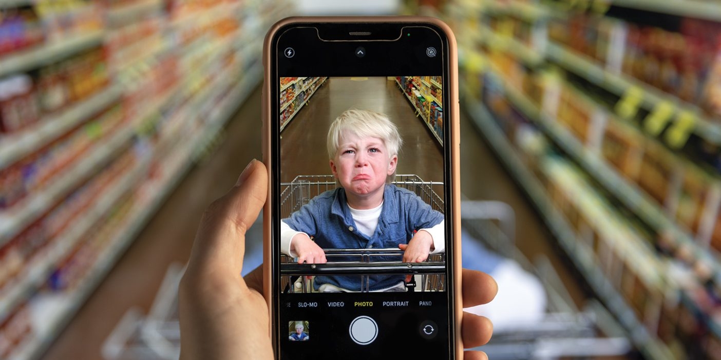 A phone zooms in on a grumpy toddler sitting in a cart in the grocery store.