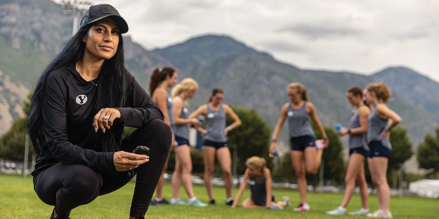 A woman in a baseball hat crouches on a sports field holding a stopwatch. Female athletes stretch in the background.