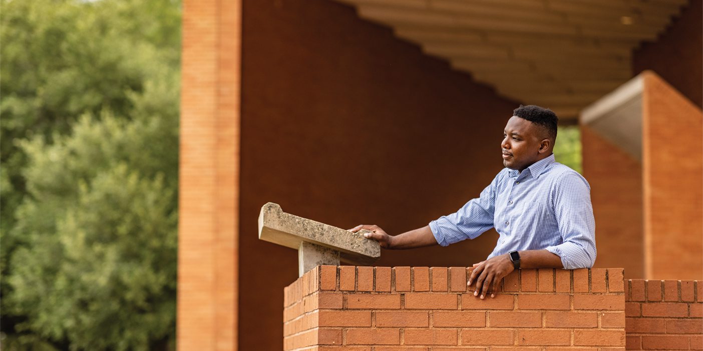 A Black man dressed in a button-down shirt stands in front of a stone podium.