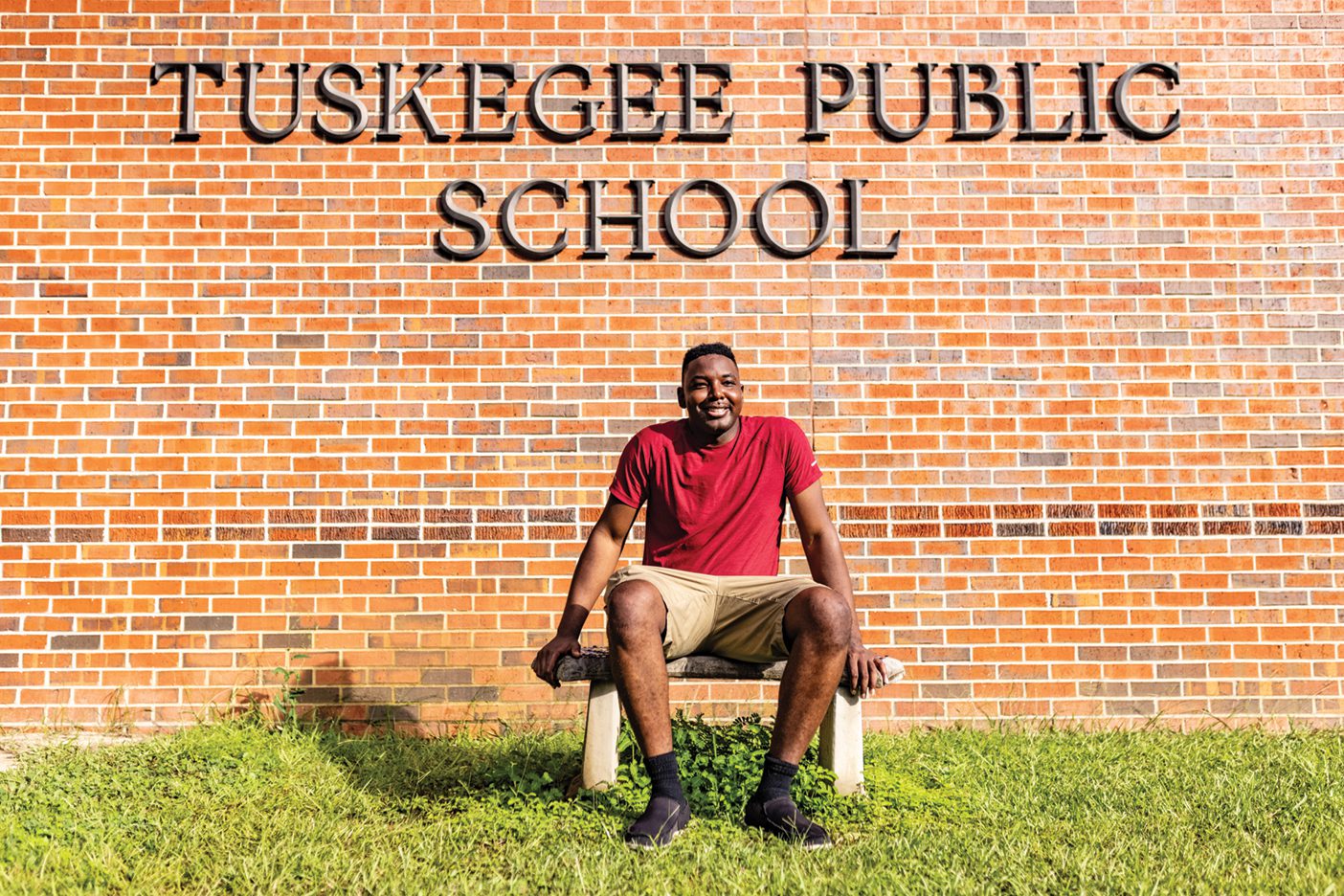 A Black man sits outside a red-brick school with engraved letters that say "Tuskegee Public School."