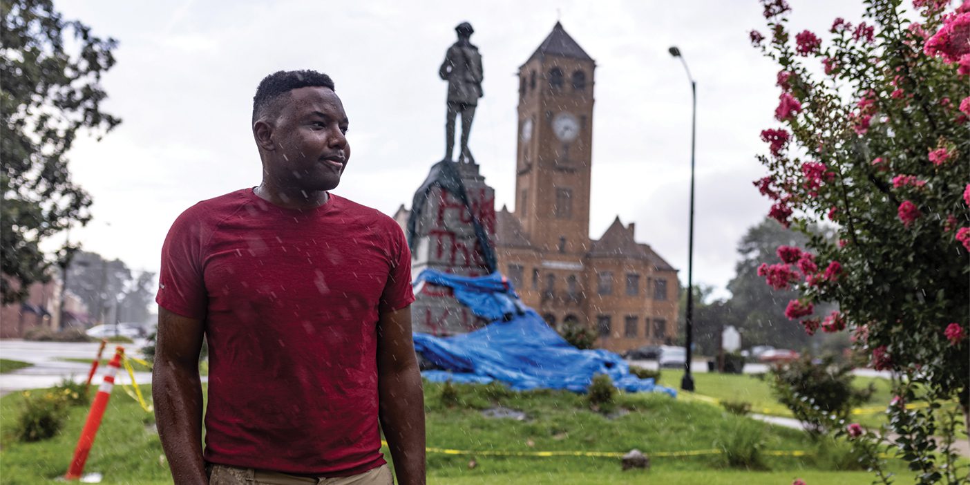 A Black man stands in a park in the rain. Behind him is a statue covered in graffiti and is marked off with caution tape.