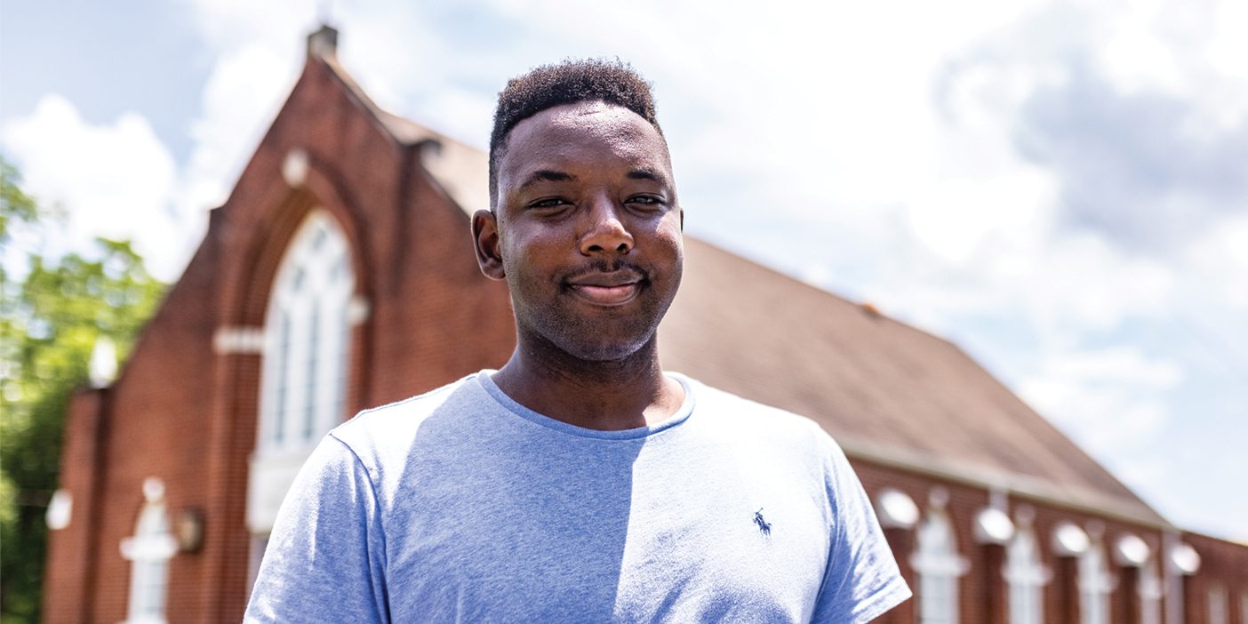 A Black man stands outside of a red-brick church.