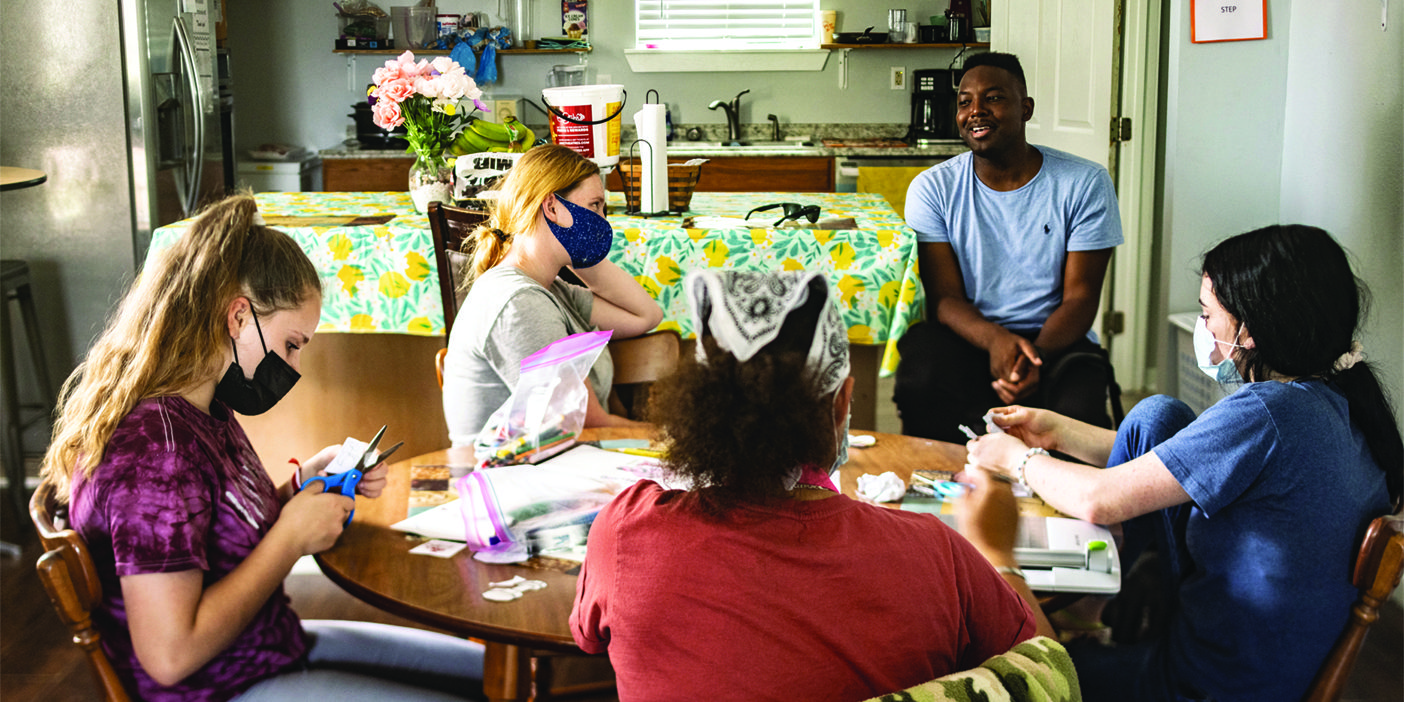 A Black man smiles and talks with a group of women coloring, cutting paper, and working on crafts.