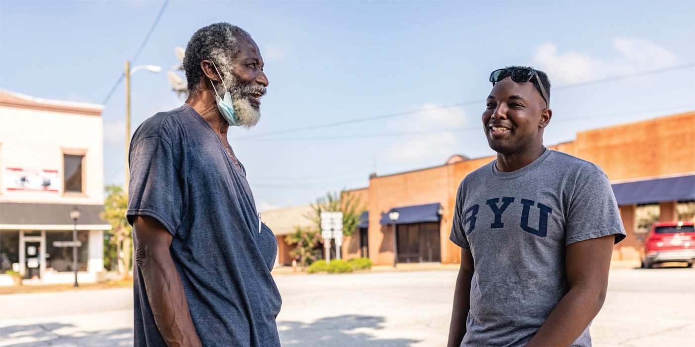 A Black man wearing a BYU shirt is talking to another Black man with a white beard and a mask.