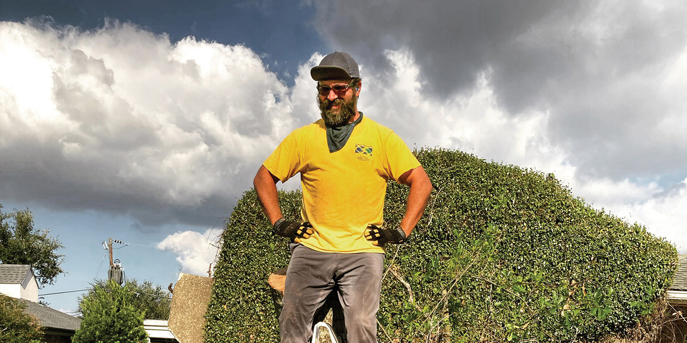 Paul Solouki wears a yellow Helping Hands shirt and stands atop a tree stump with a chainsaw.