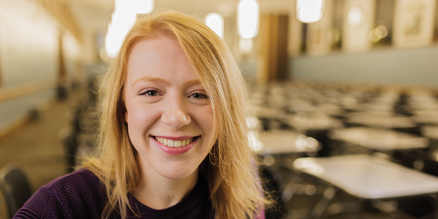 A young woman smiles the the testing center, an empty room full of desks and chairs.