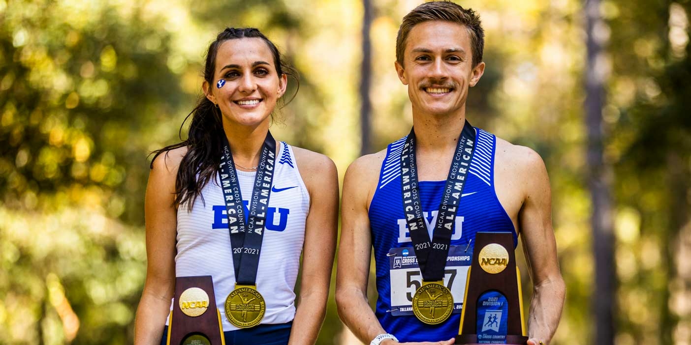 Female and male runners in BYU uniforms stand next to each other wearing first-place medals and holding trophies.