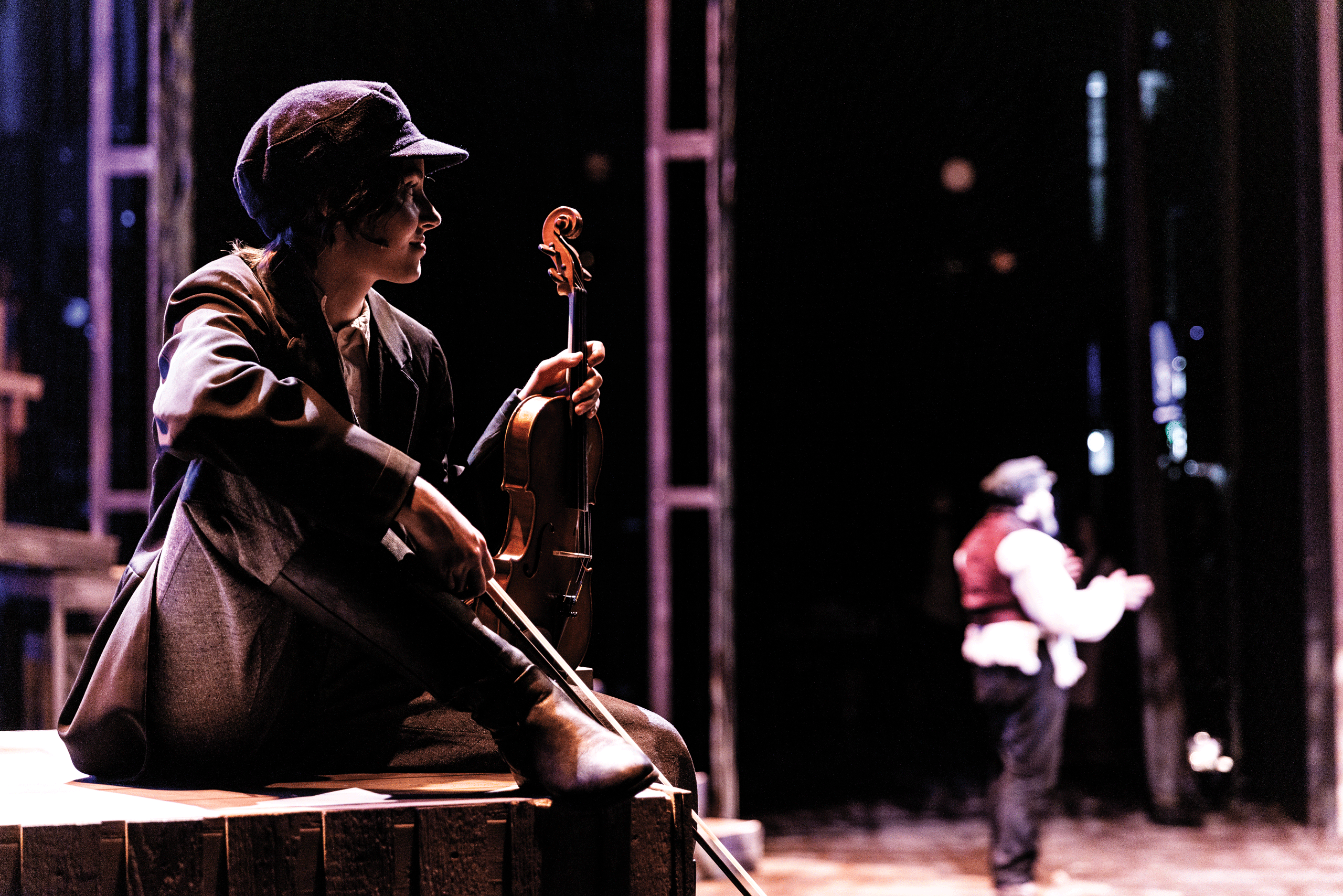 On a stage during a live performance of the Fiddler on the Roof in BYU's de Jong Concert Hall, a female student sits in the foreground holding a violin, gazing at an actor in the background speaking to an audience