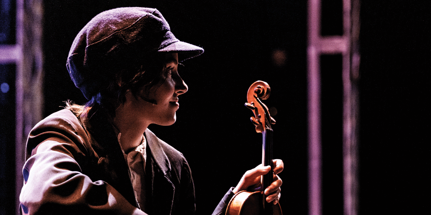 On a stage during a live performance of the Fiddler on the Roof, a female student sits in the foreground holding a violin, gazing at an actor in the background speaking to an audience