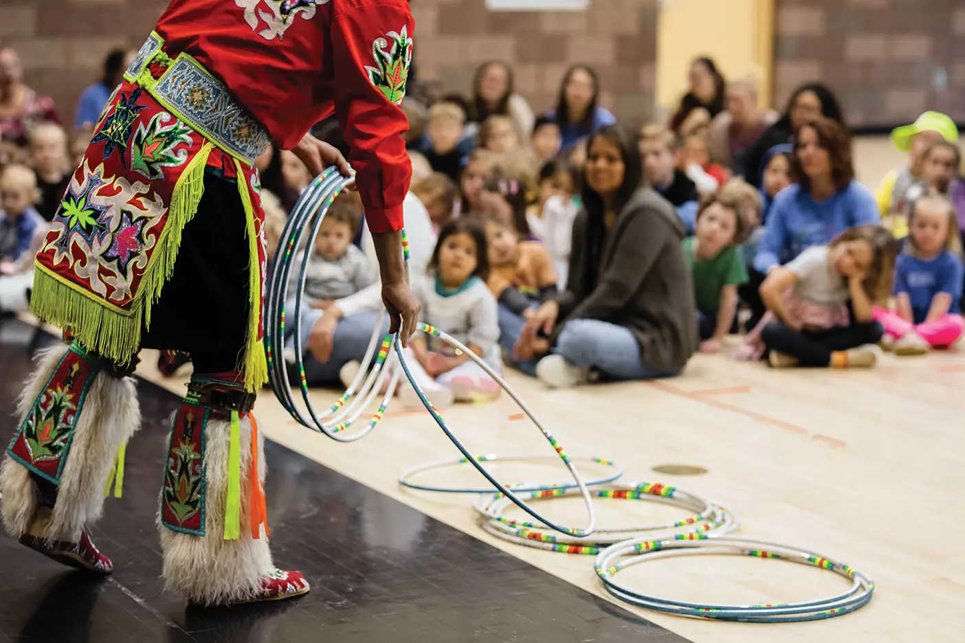 An Indigenous dancer, dressed in traditional clothing, places beaded hoops on the gym floor as elementary school children sit and watch.