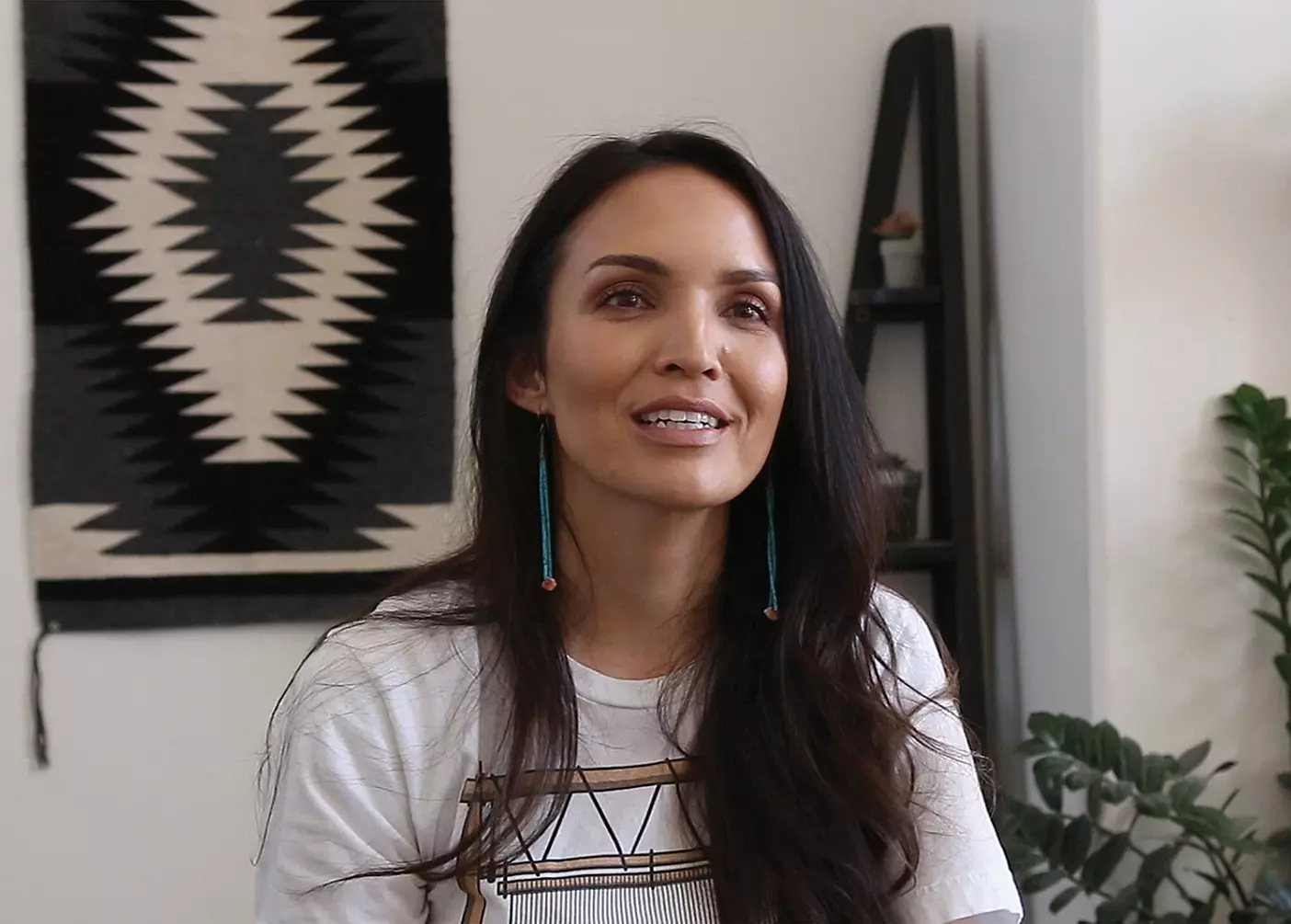 Michele Reyes sits in her office, talking to an interviewer behind the camera. A black and white piece of geometric woven art is hangs on the wall behind her.