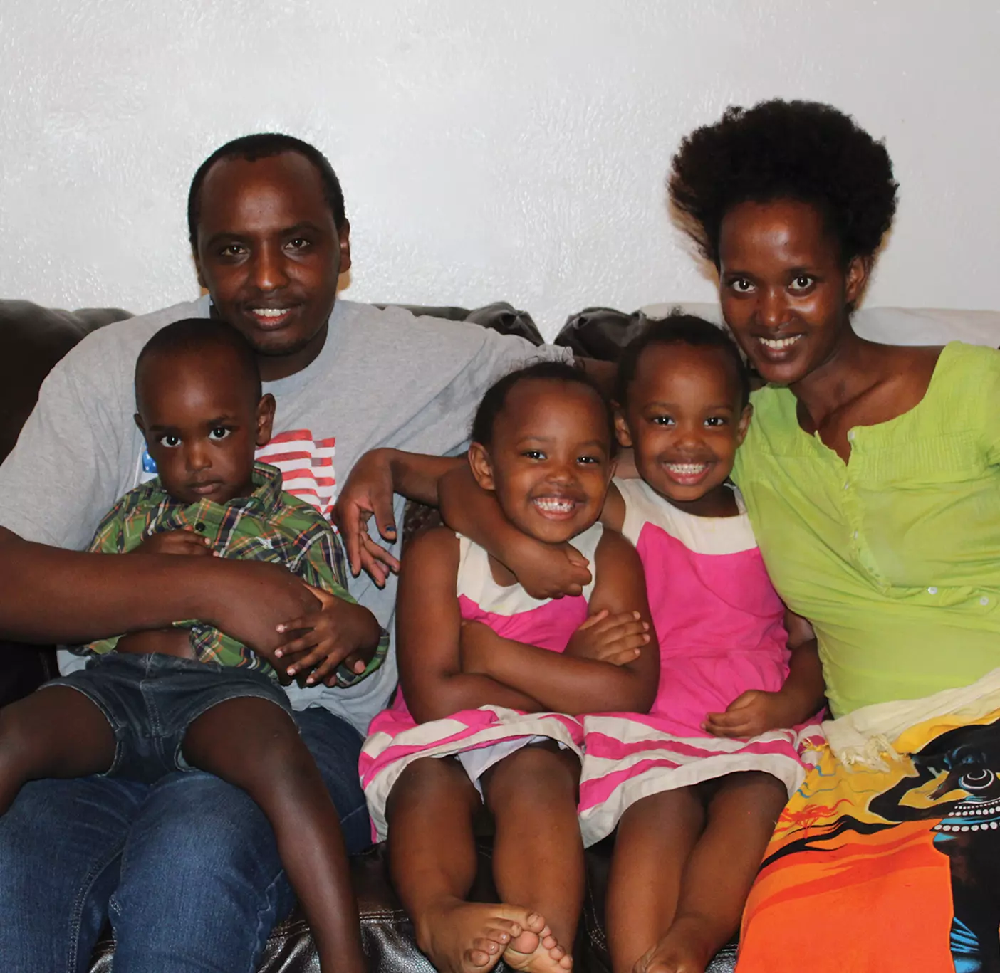 A refugee family with three young children smiles and cuddles on the couch.