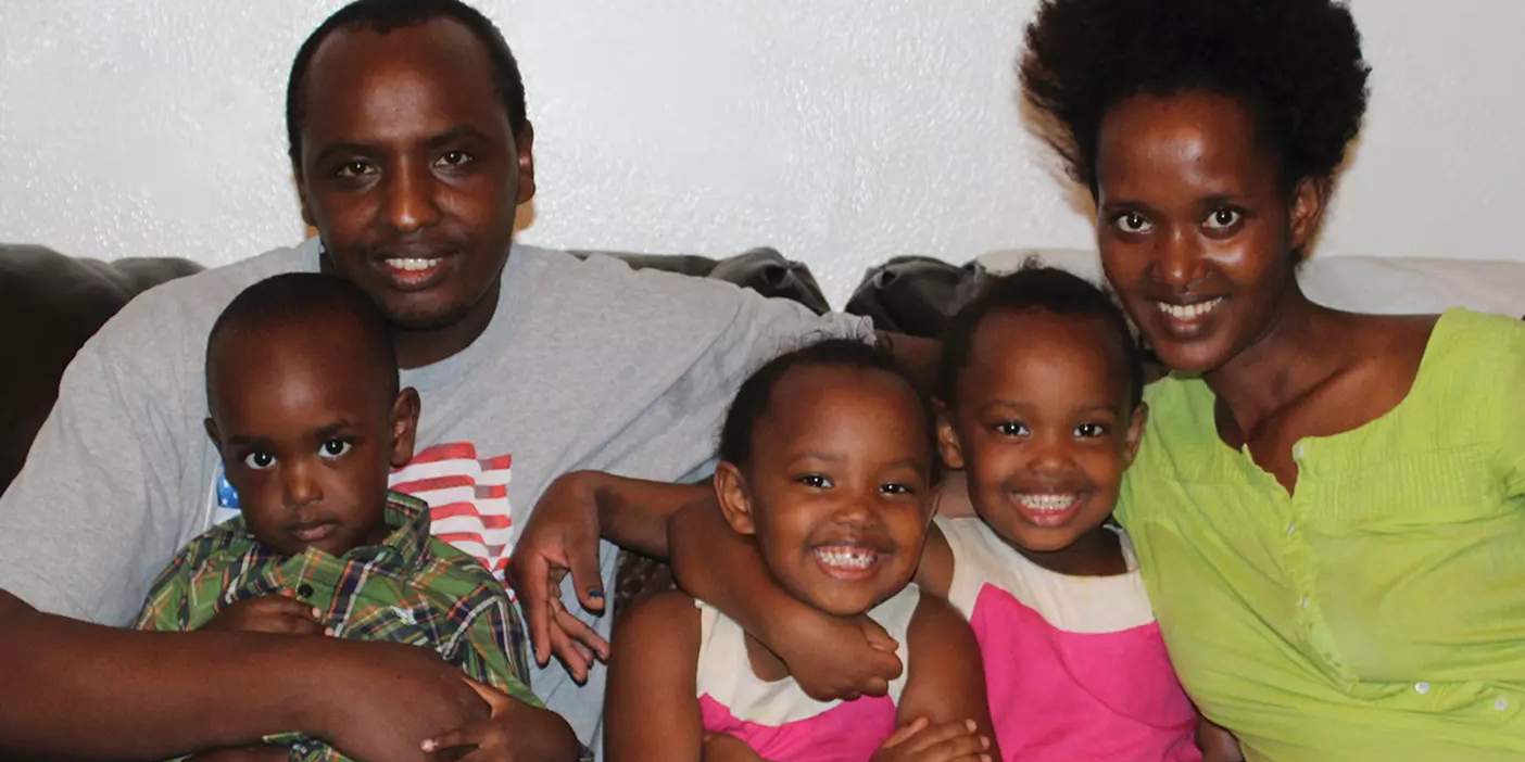 A refugee family with three young children smiles and cuddles on the couch.