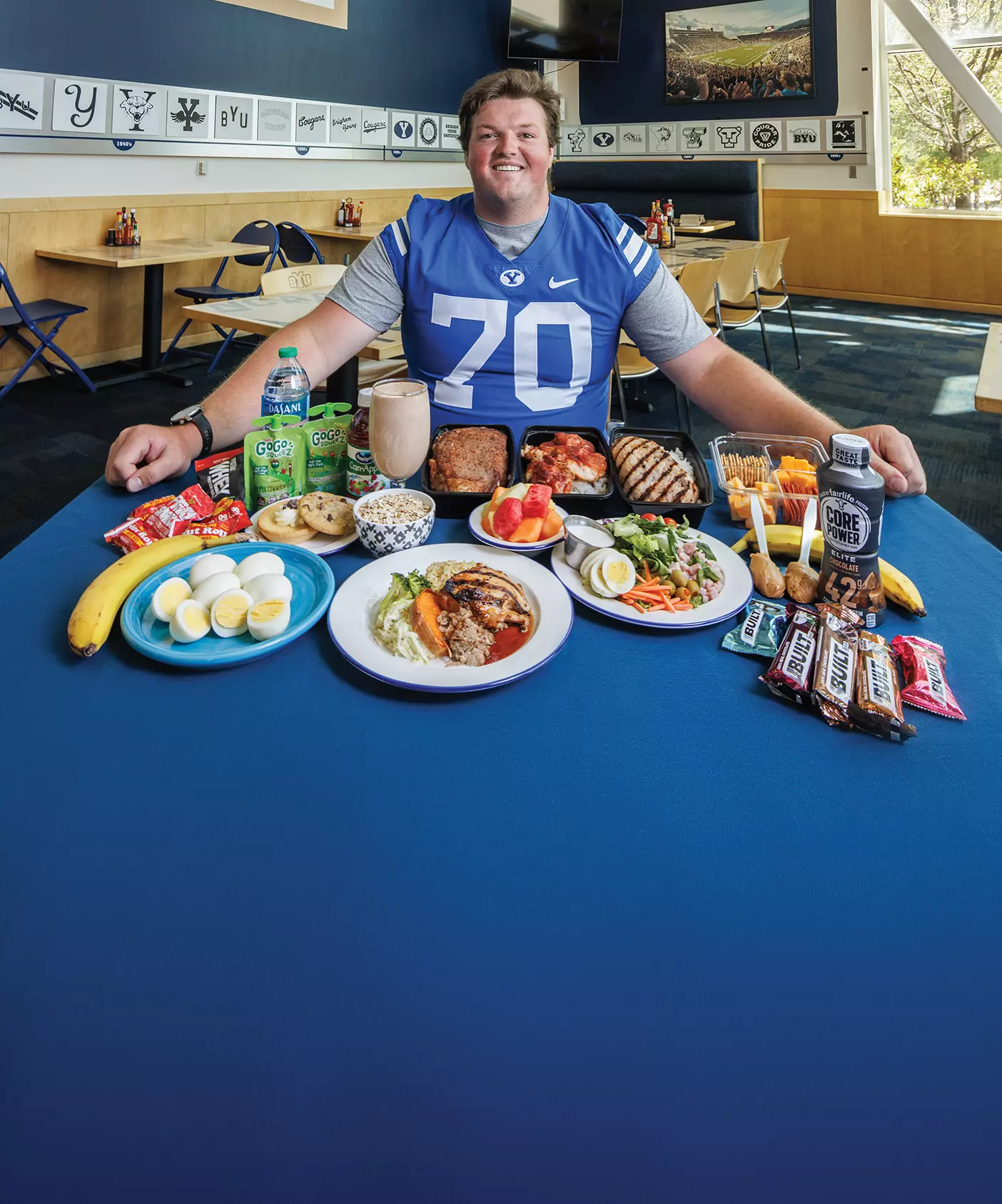 A football player is surrounded by the food he eats in a day, including eggs, oatmeal, smoothie, meat, fruit, and protein bars.