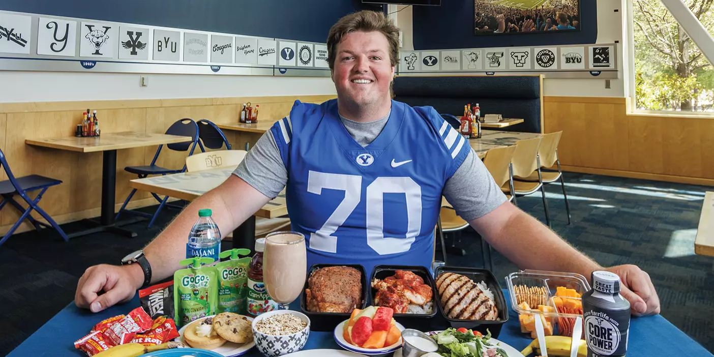 A football player is surrounded by the food he eats in a day, including eggs, oatmeal, smoothie, meat, fruit, and protein bars.