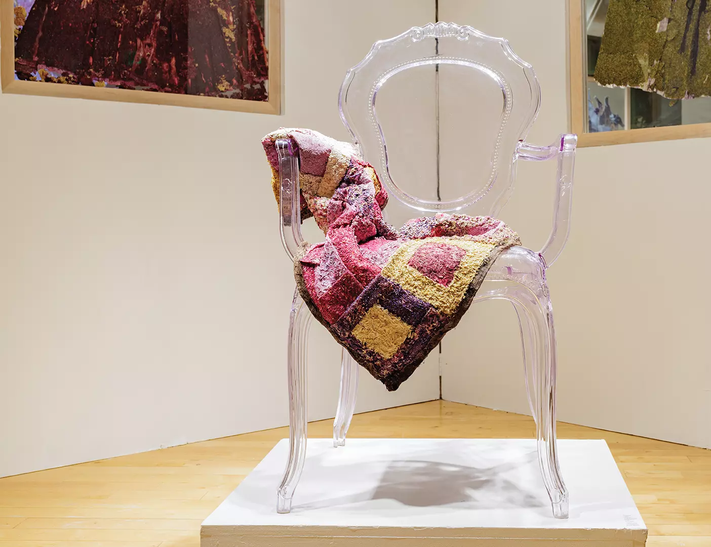 A quilt made from dried flowers is draped over a clear chair in an art gallery