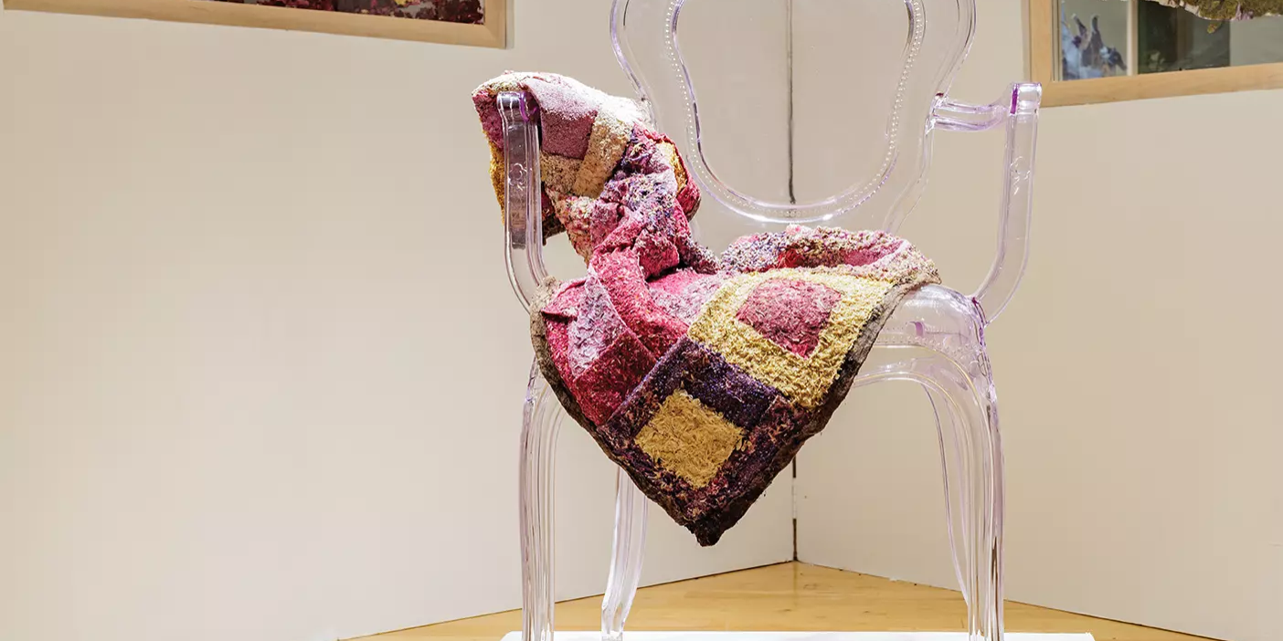 A quilt made from dried flowers is draped over a clear chair in an art gallery