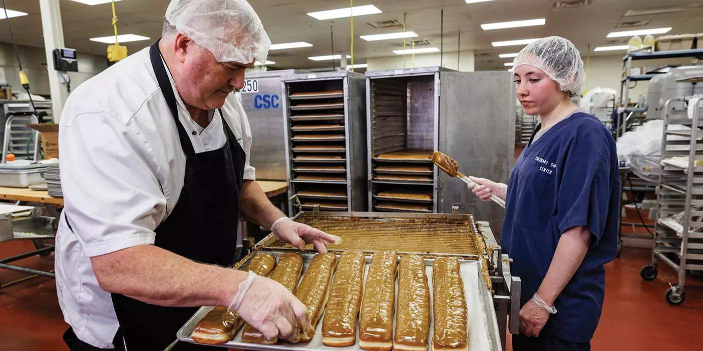 Two dining services employees wearing hair nets frost a line of 15-inch long Cougar Tails, a huge maple bar treat.