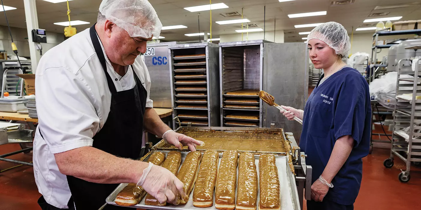 Two dining services employees wearing hair nets frost a line of 15-inch long maple bars.