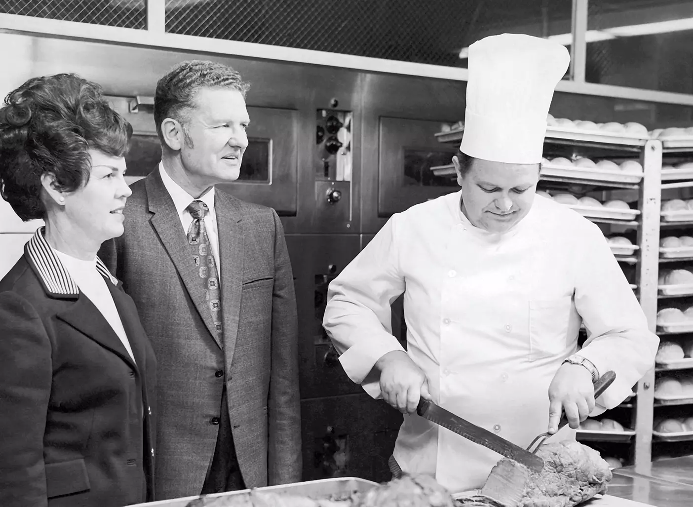 A black-and-white photo of Wells and Myrle Cloward supervising a kitchen and a chef cutting roast.