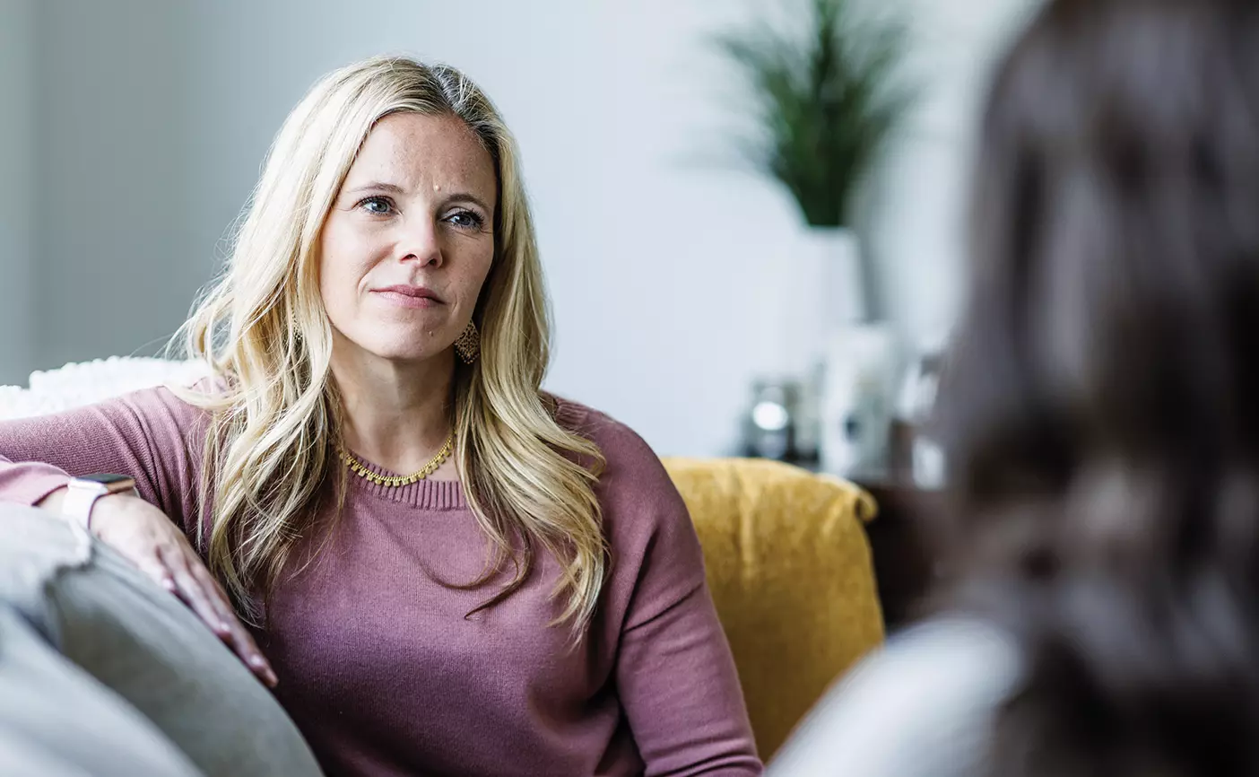 A therapist in a pink sweater sits on a couch and counsels another woman.