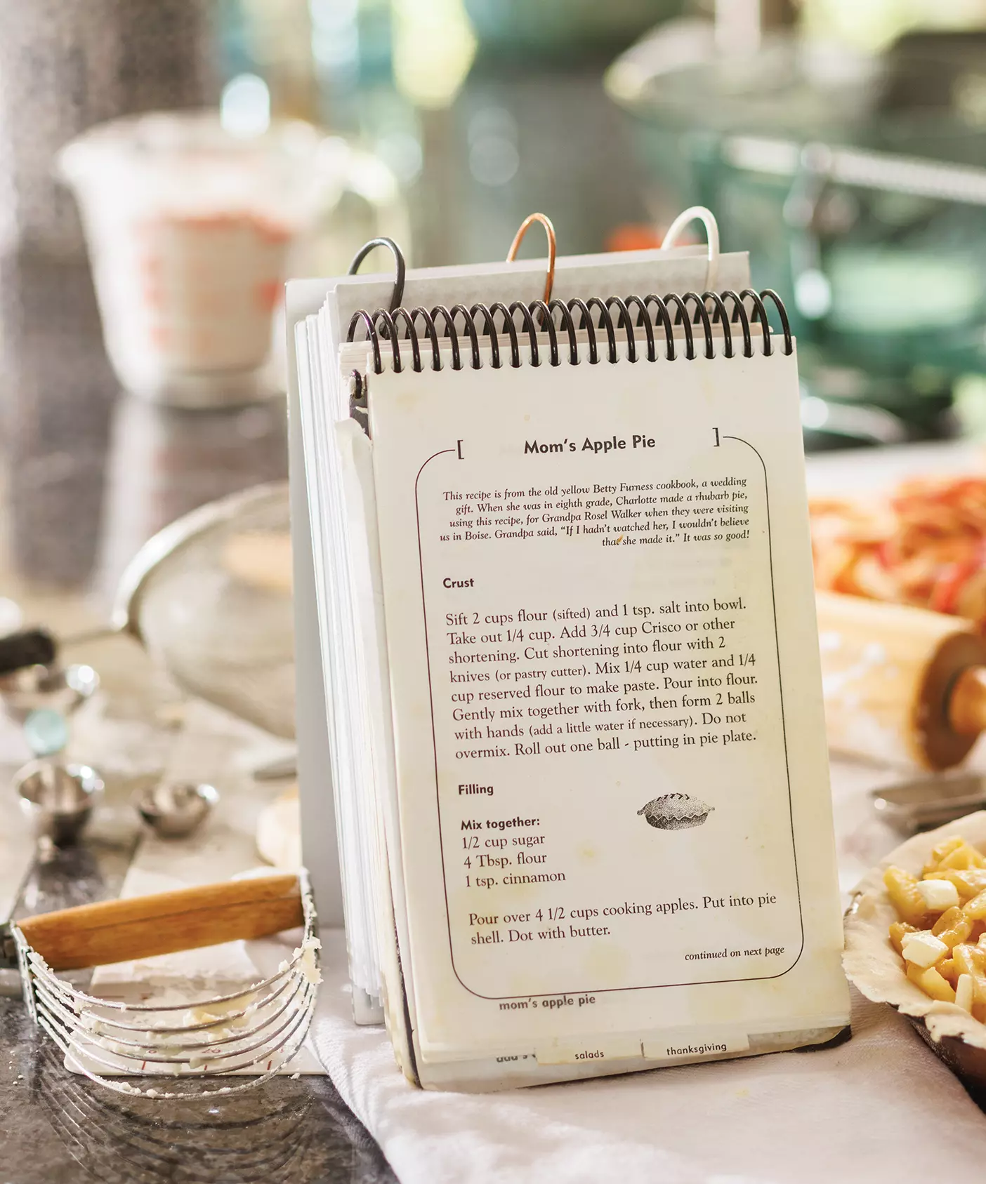 A homemade cookbook opened up to a family apple pie recipe sits next to a pastry cutter, measuring spoons, a sifter, a rolling pin, apple peels, and a half-constructed pie dotted with butter.