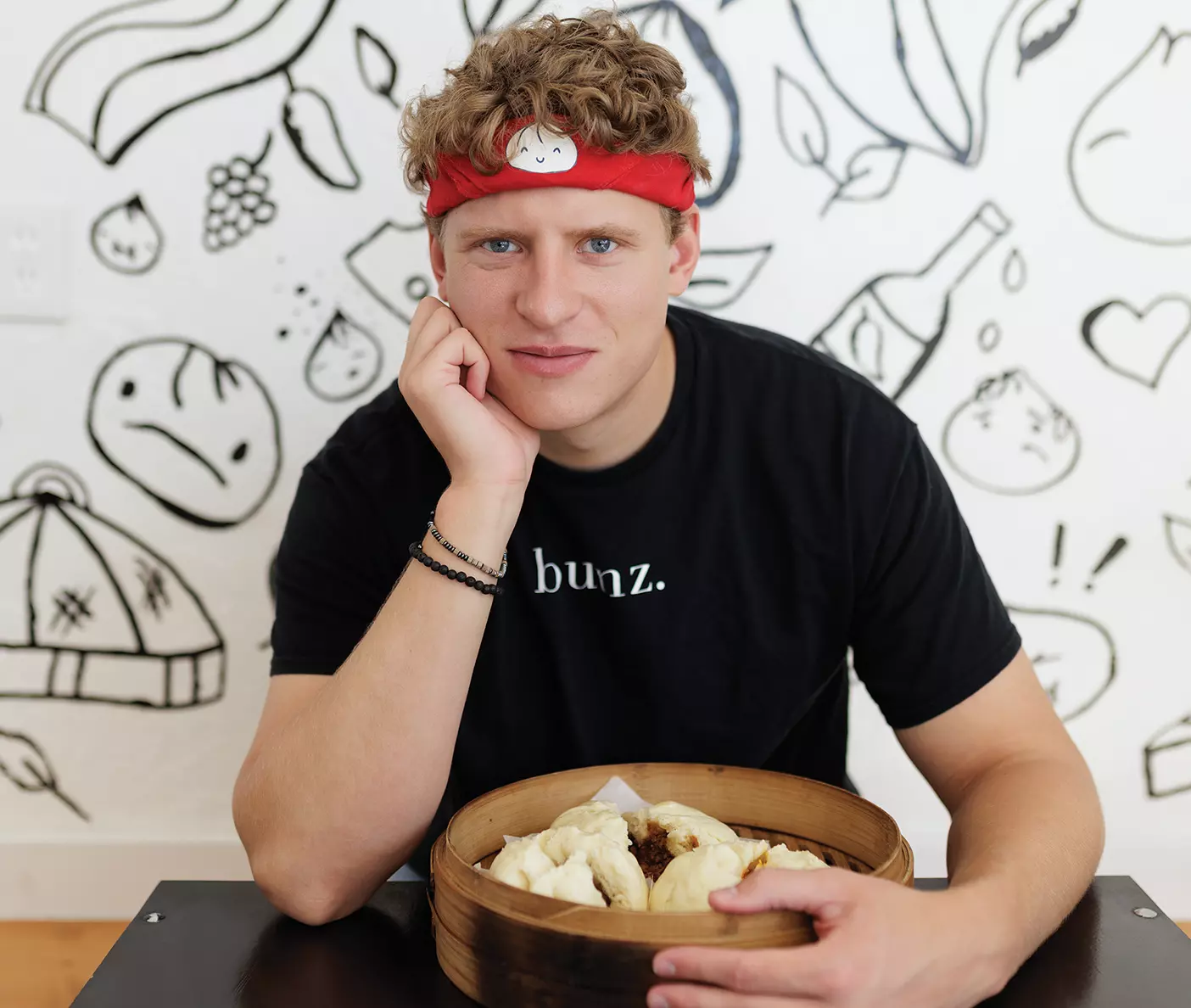 Jimmy Watson sits at Bun Boiz with a basket of steamed buns in front of him. He wears a red headband with a dumpling on it and a black Bun Boiz shirt.