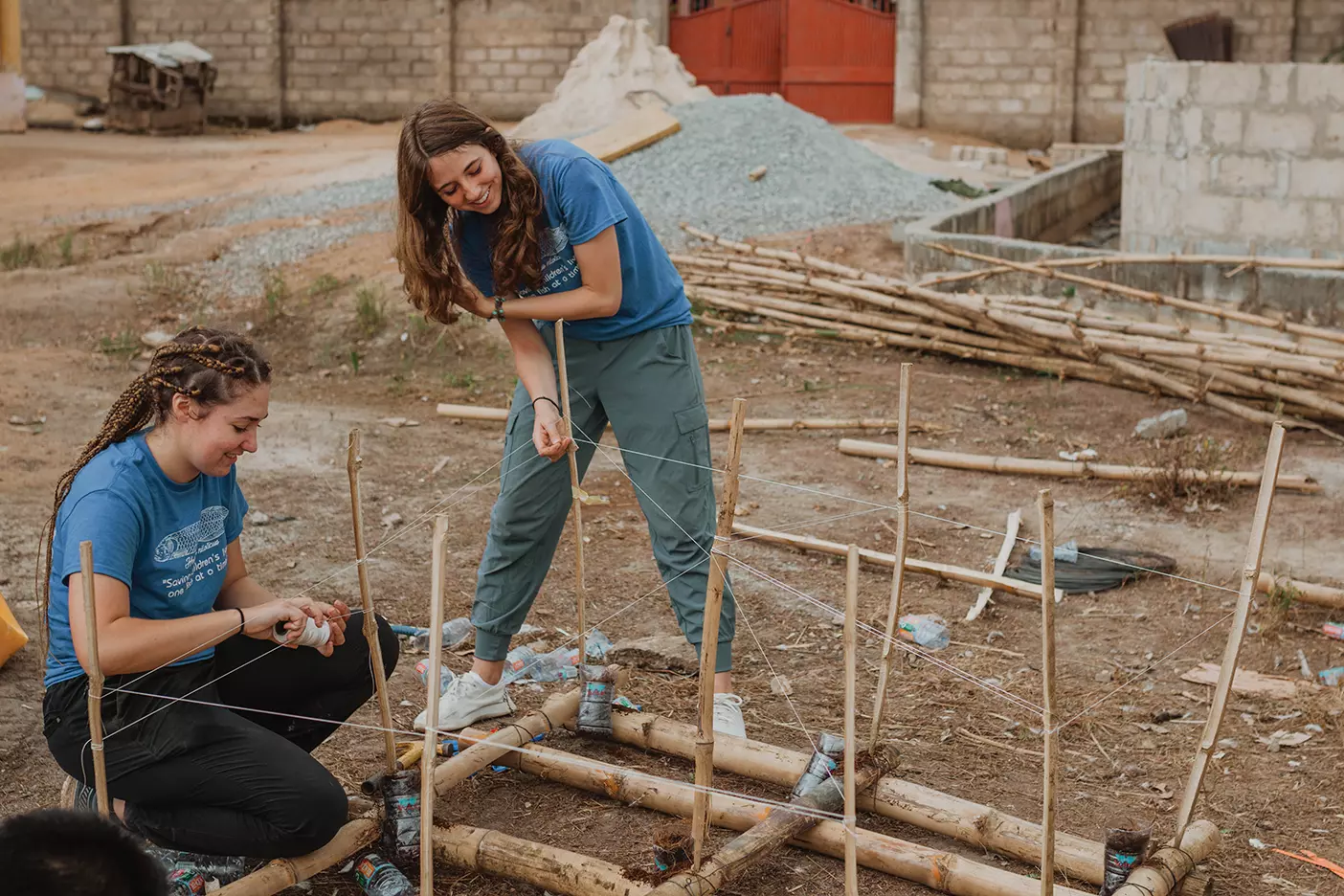 Two women in blue shirts construct a raft out of bamboo. They are working outside in a patch of dirt. 