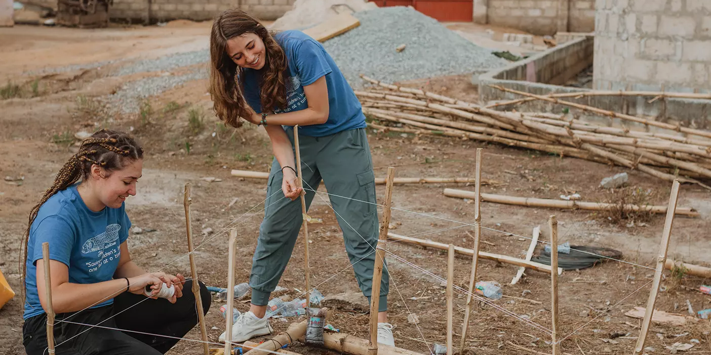 Two women in blue shirts construct a raft out of bamboo. They are working outside in a patch of dirt.