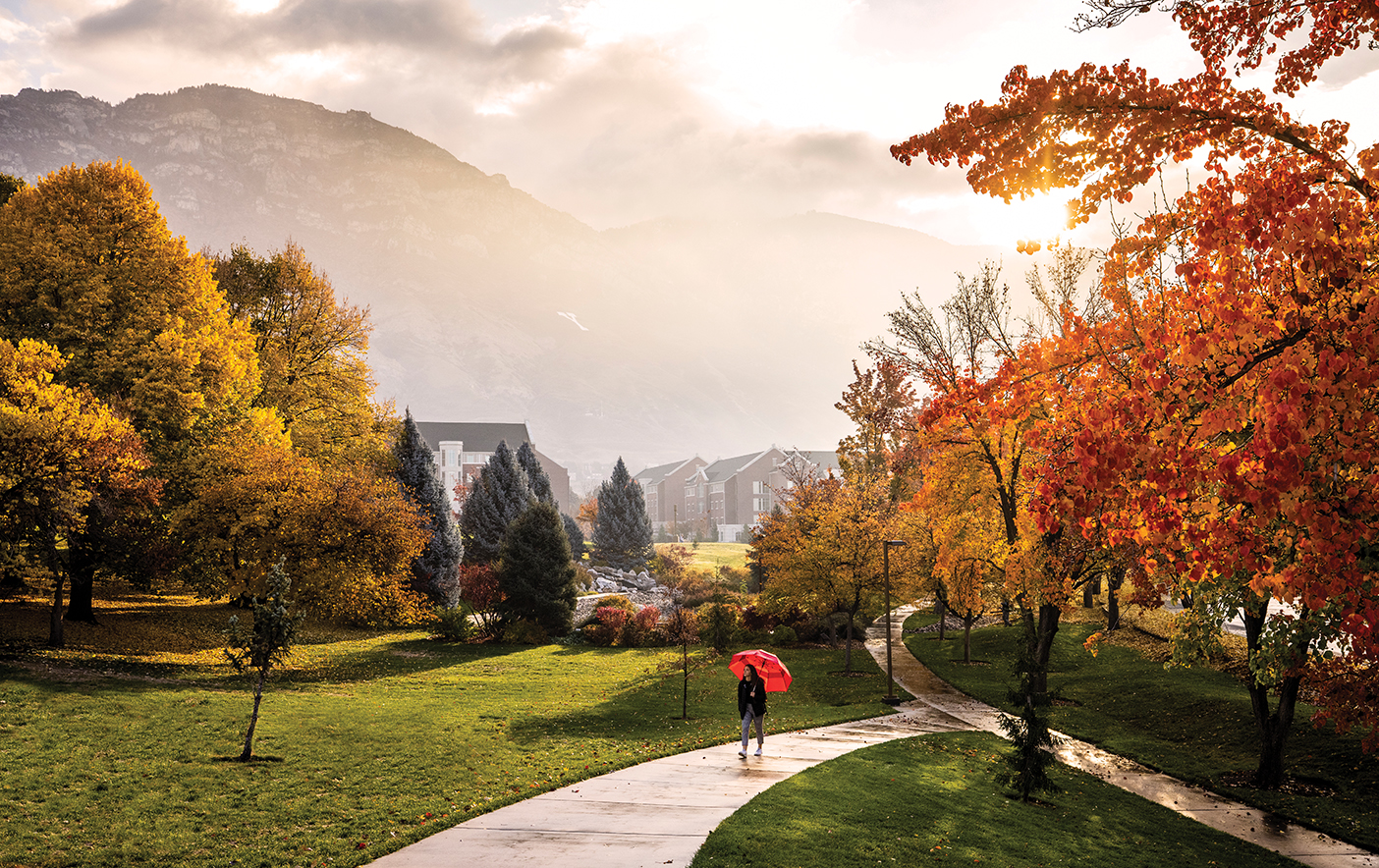 A student walks on BYU campus carrying a red umbrella. The mountains behind her are foggy, and the photo is framed by trees with red and yellow leaves.