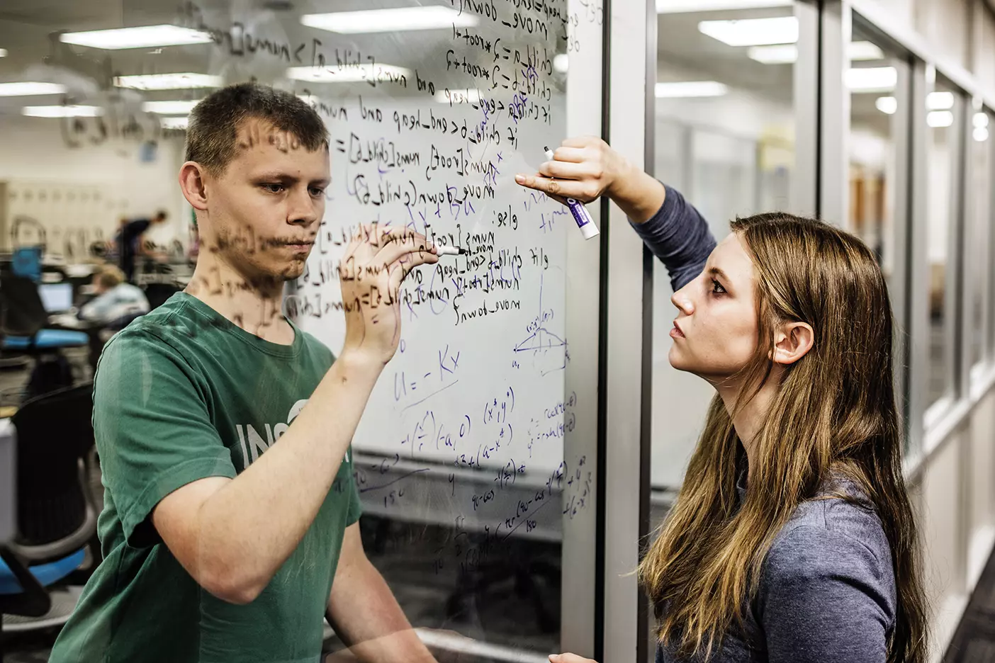 Two students, each on opposite sides of an indoor glass window, write math equations together on the window. 