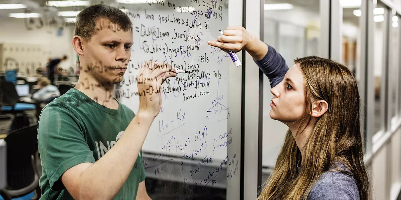 Two students, each on opposite sides of an indoor glass window, write math equations together on the window.