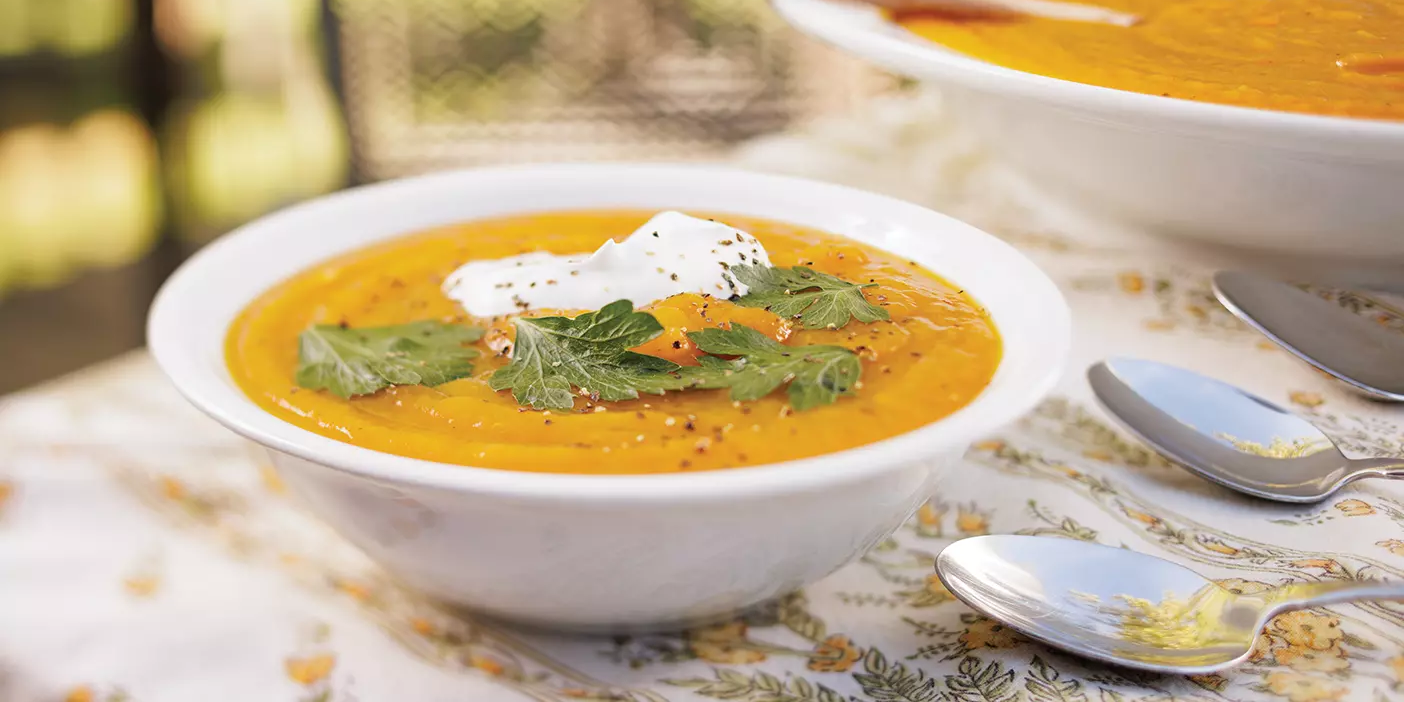 Orange pumpkin soup topped with herbs and cream is served in a white bowl.