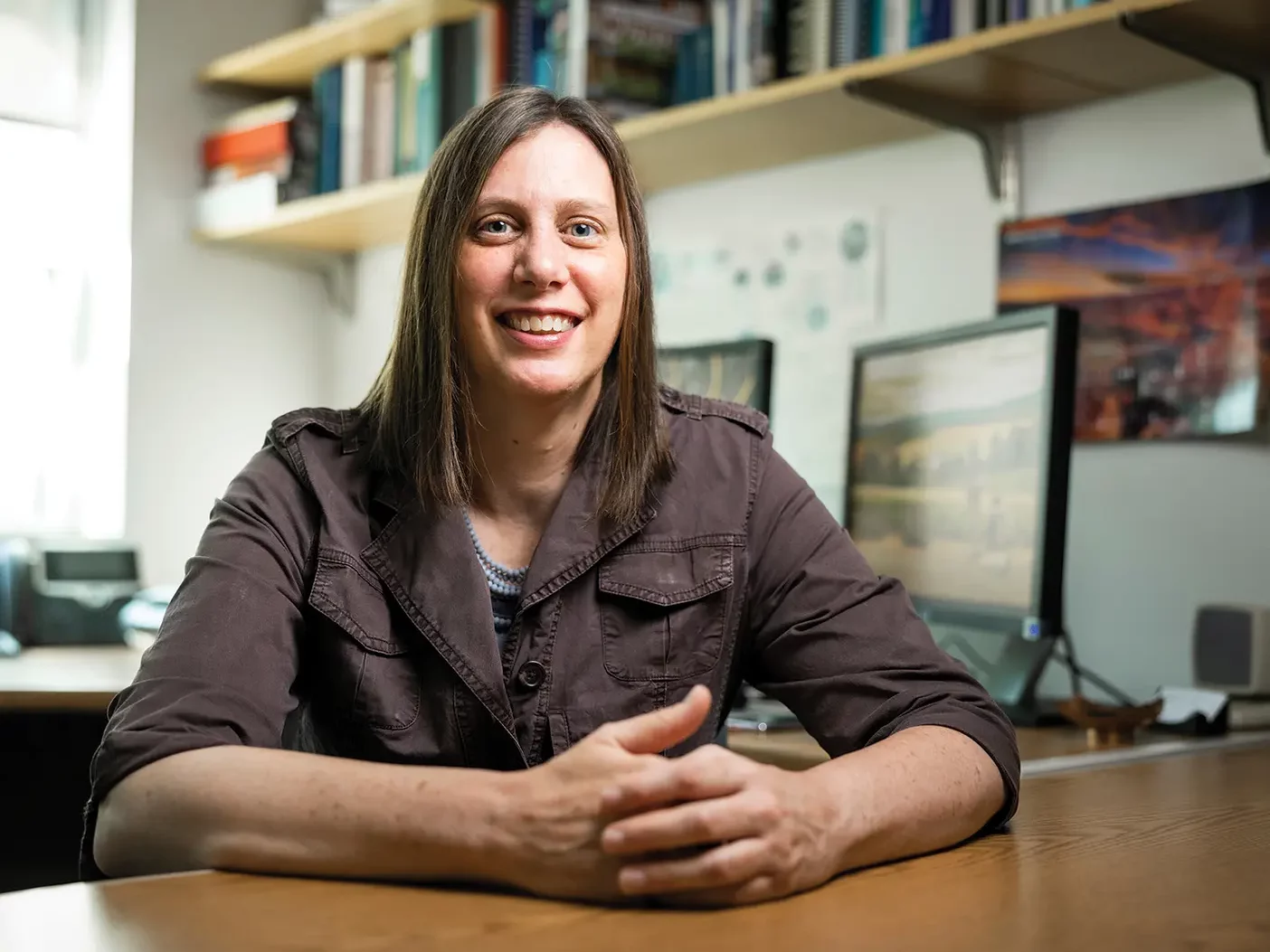 A female professor with brown hair and light skin sits in her office, surrounded by books and computer screens, smiling at the camera