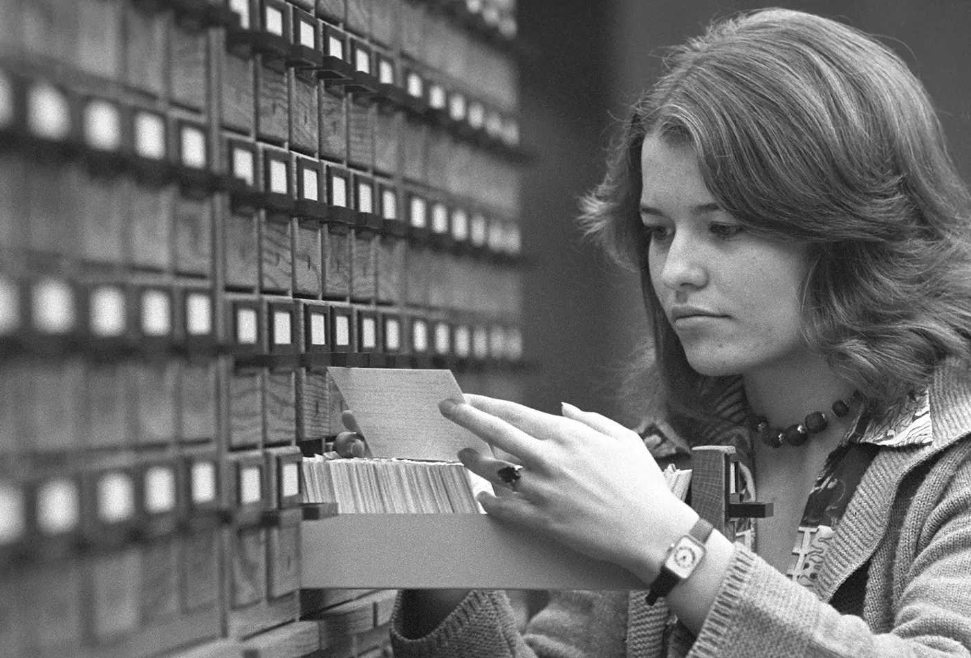 A black-and-white image of a female student looking up something in a card catalog.