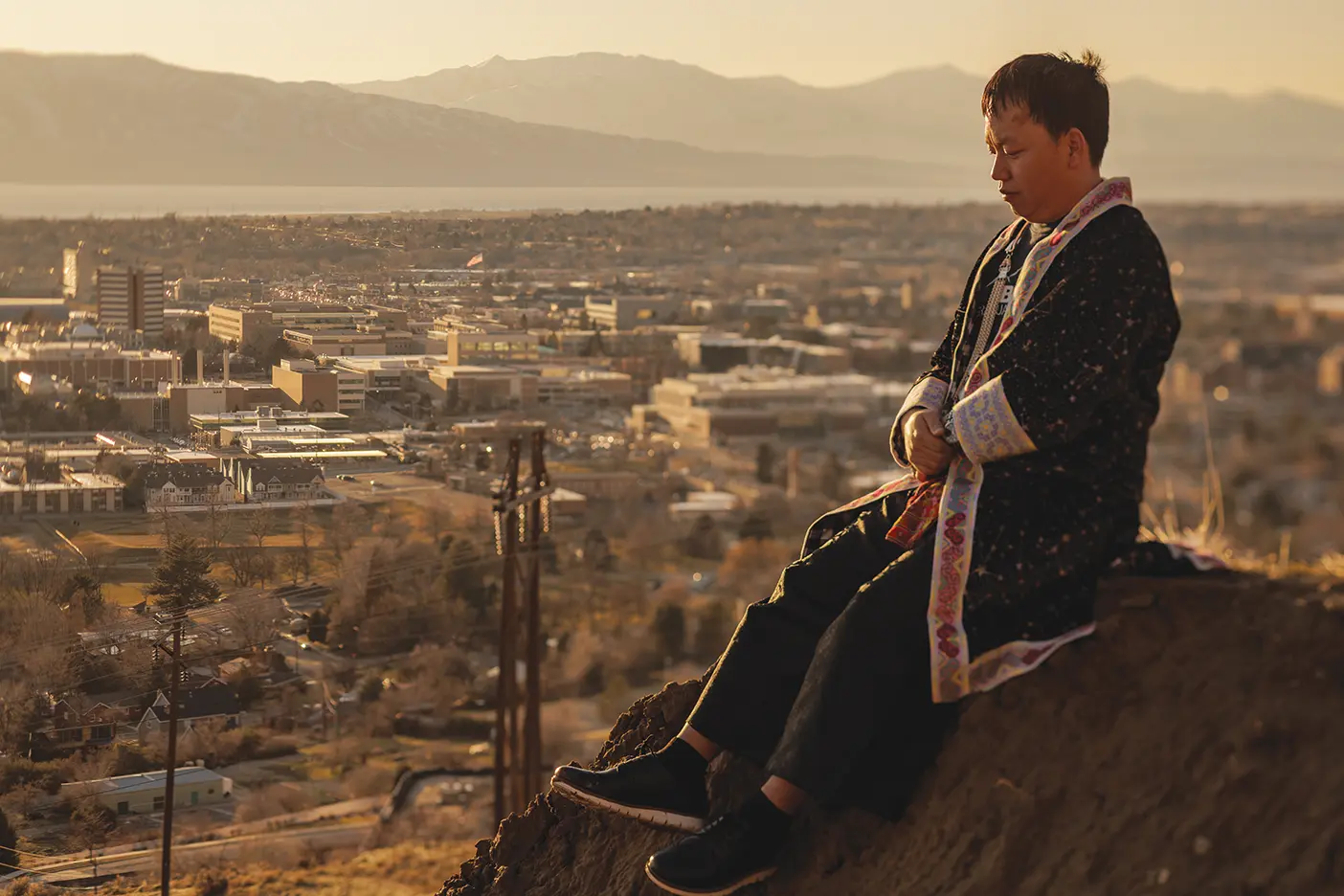 Yang Vang sits on a sun-bathed slope overlooking BYU's campus, wearing traditionall Hmong clothing.